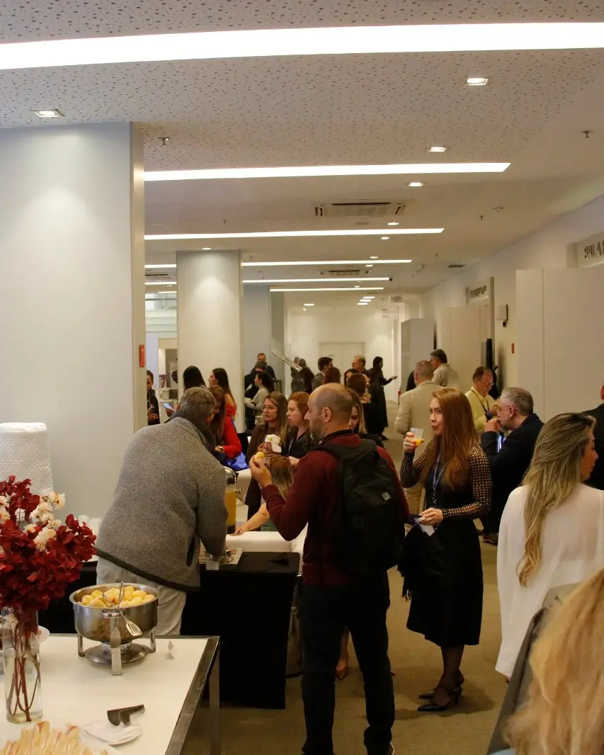 Participantes de pé conversando durante o coffee break, com os stands da feira ao fundo, em uma edição anterior.